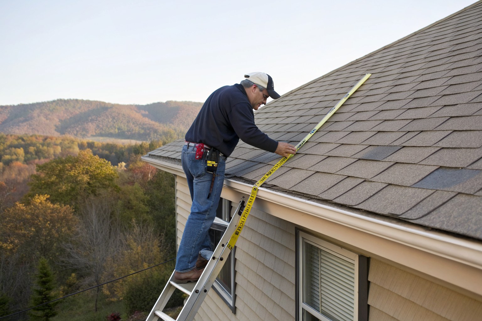 A man on a ladder measuring a shingle roof with a tape, with wooded hills behind