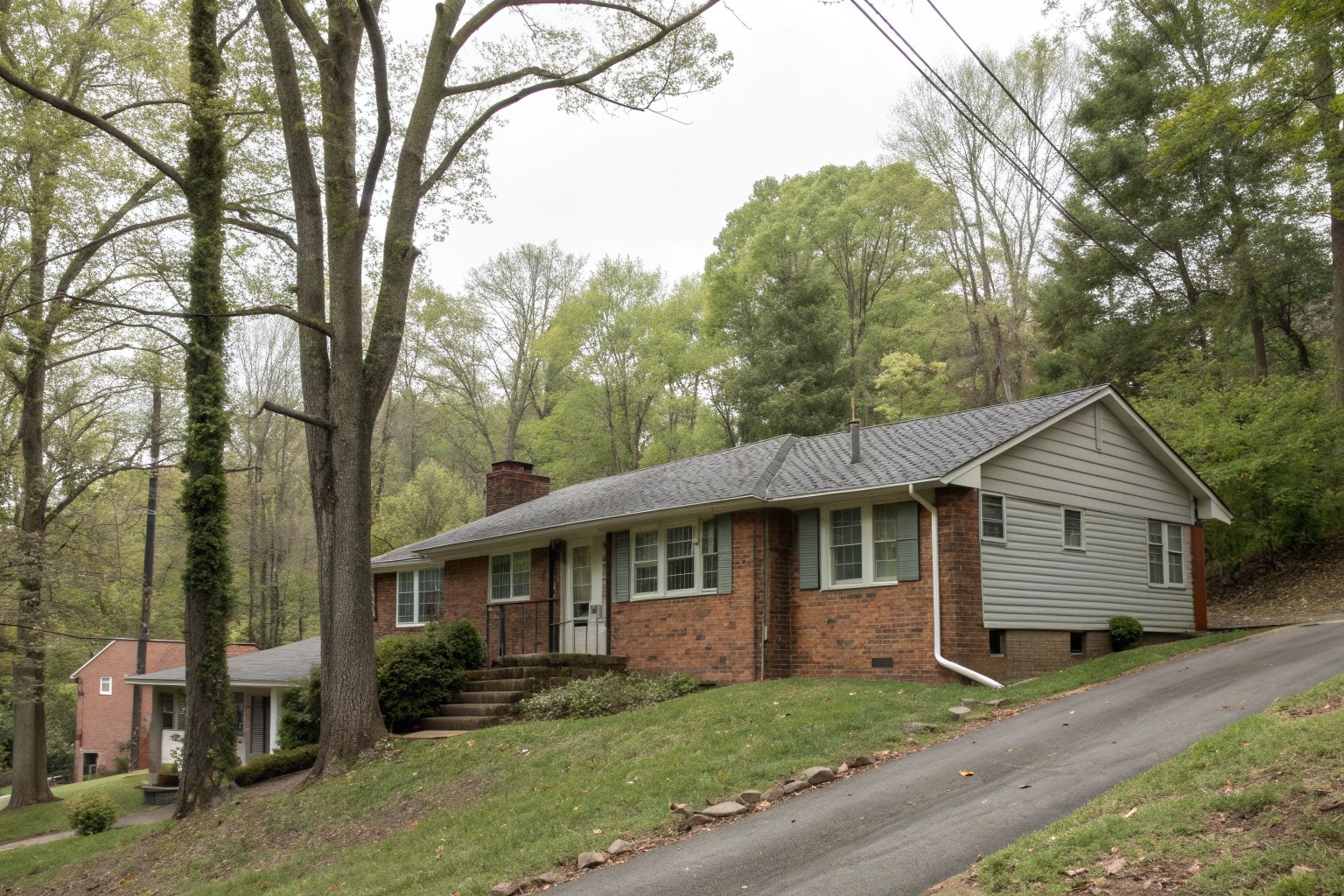 A brick and siding ranch house with a gray shingle roof among tall trees